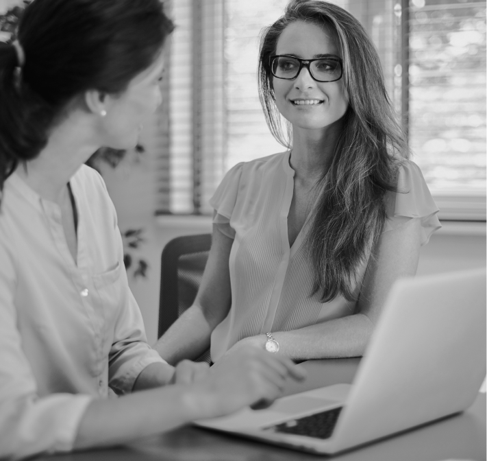 Duas mulheres estão sentadas à mesa com um laptop e sorrindo